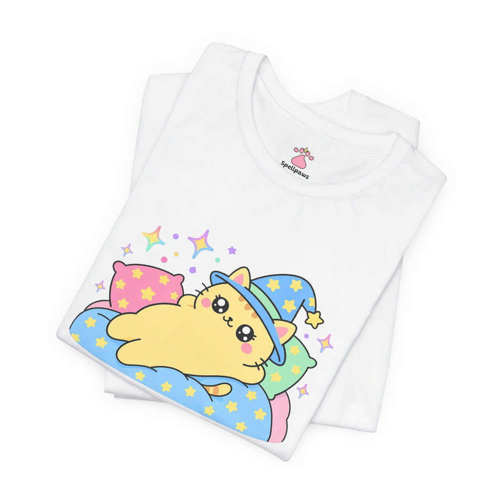 Folded white Pillowus Conquestus T-shirt showing a close-up of the yellow wizard cat in a blue hat sprawled across a pile of pillows on a starry blanket