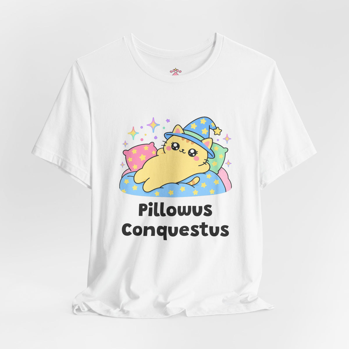 White Pillowus Conquestus wizard cat T-shirt shown in a soft studio setting: yellow wizard cat in a blue hat sprawled across a pile of pillows on a starry blanket